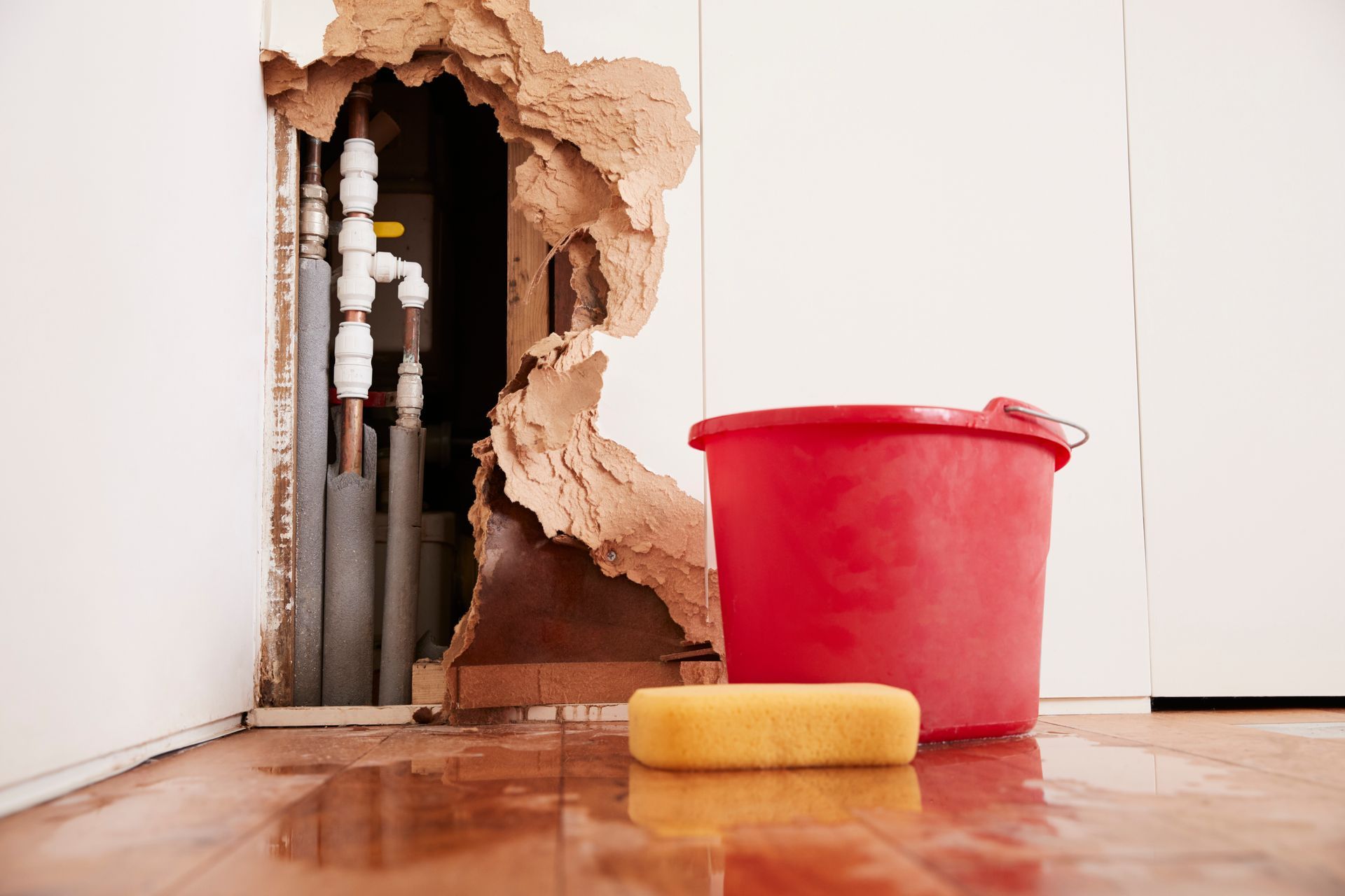 Broken wall with exposed pipes, red bucket, and yellow paint roller on a wet floor