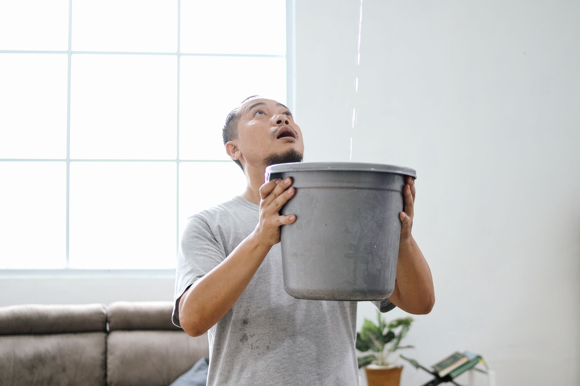 Man holding bucket, catching water leaking from ceiling. Interior room with window and sofa.