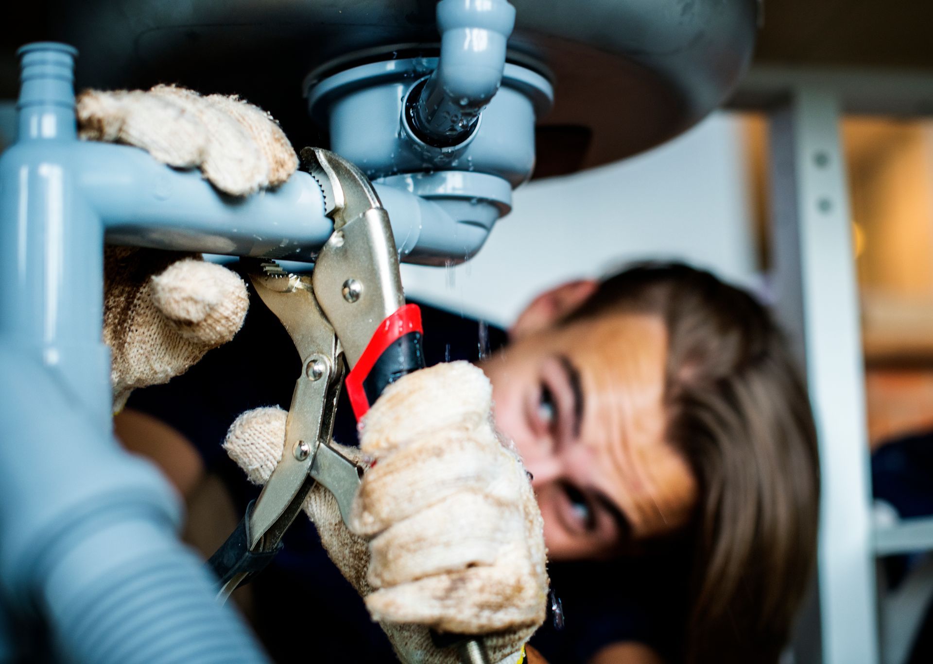 Person with gloves fixing a leaky pipe under a sink with a wrench.
