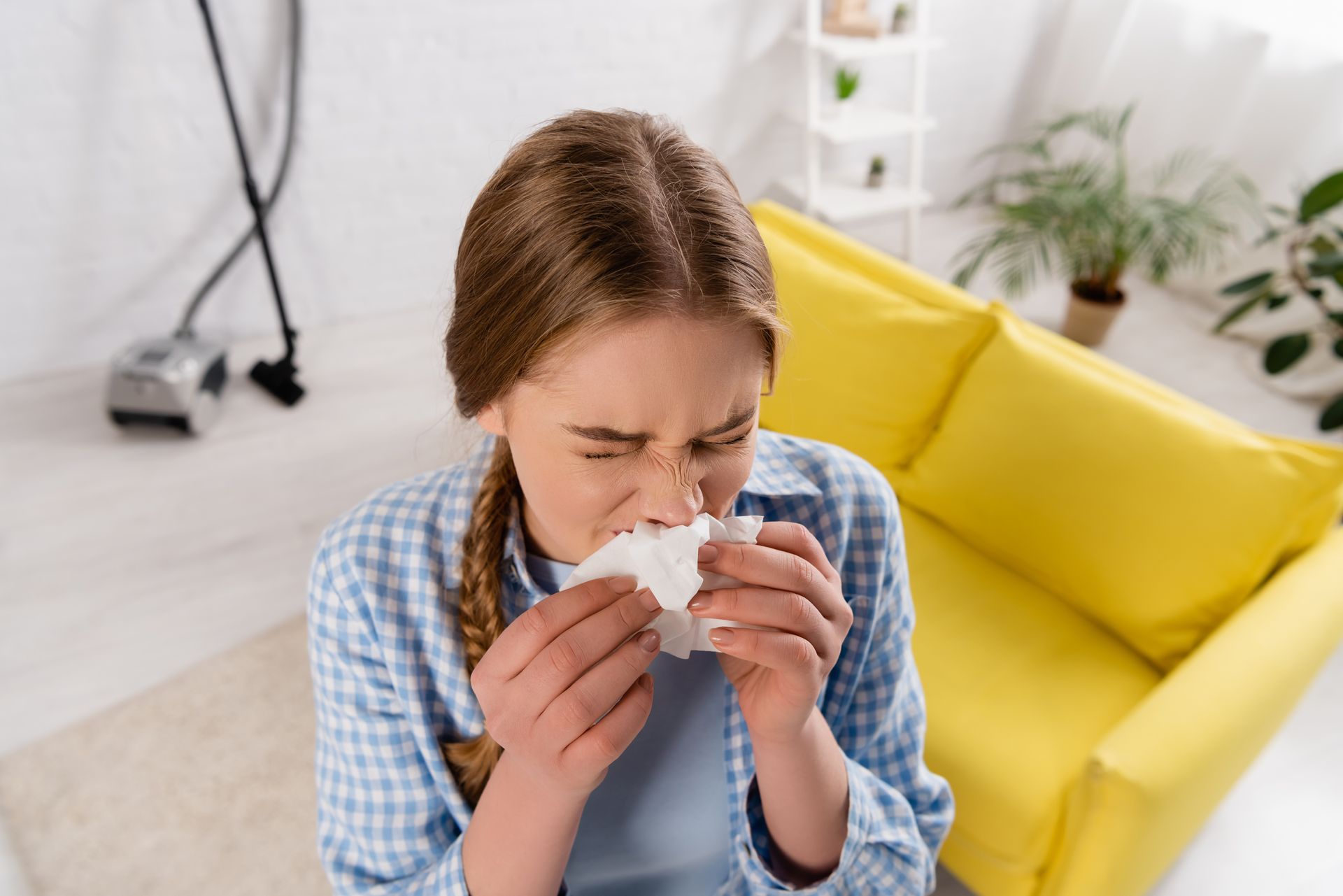 Woman sneezing into a tissue on a yellow couch in a bright living room