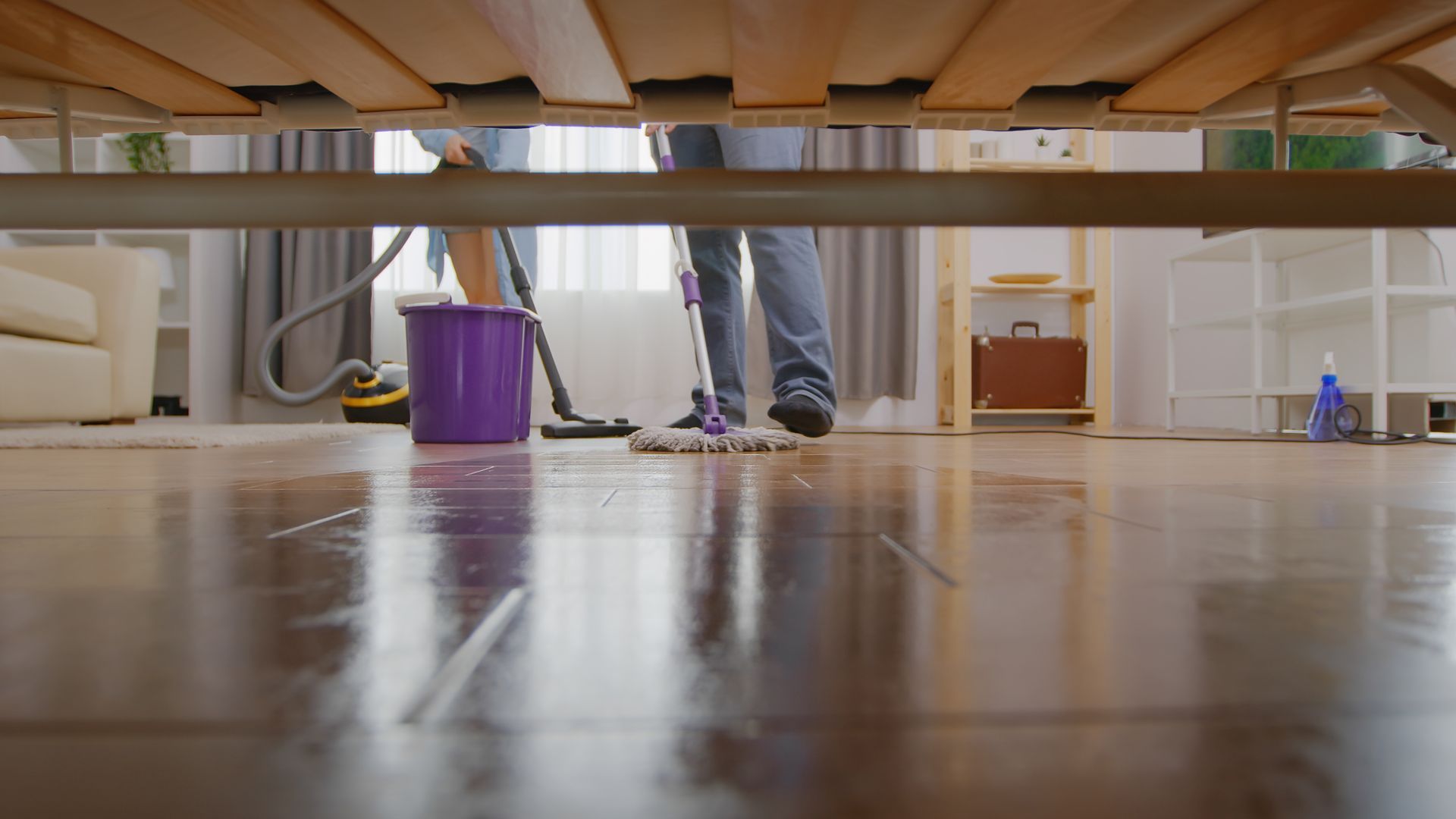 Person mopping a reflective floor under a bed, with a purple bucket in a bedroom.
