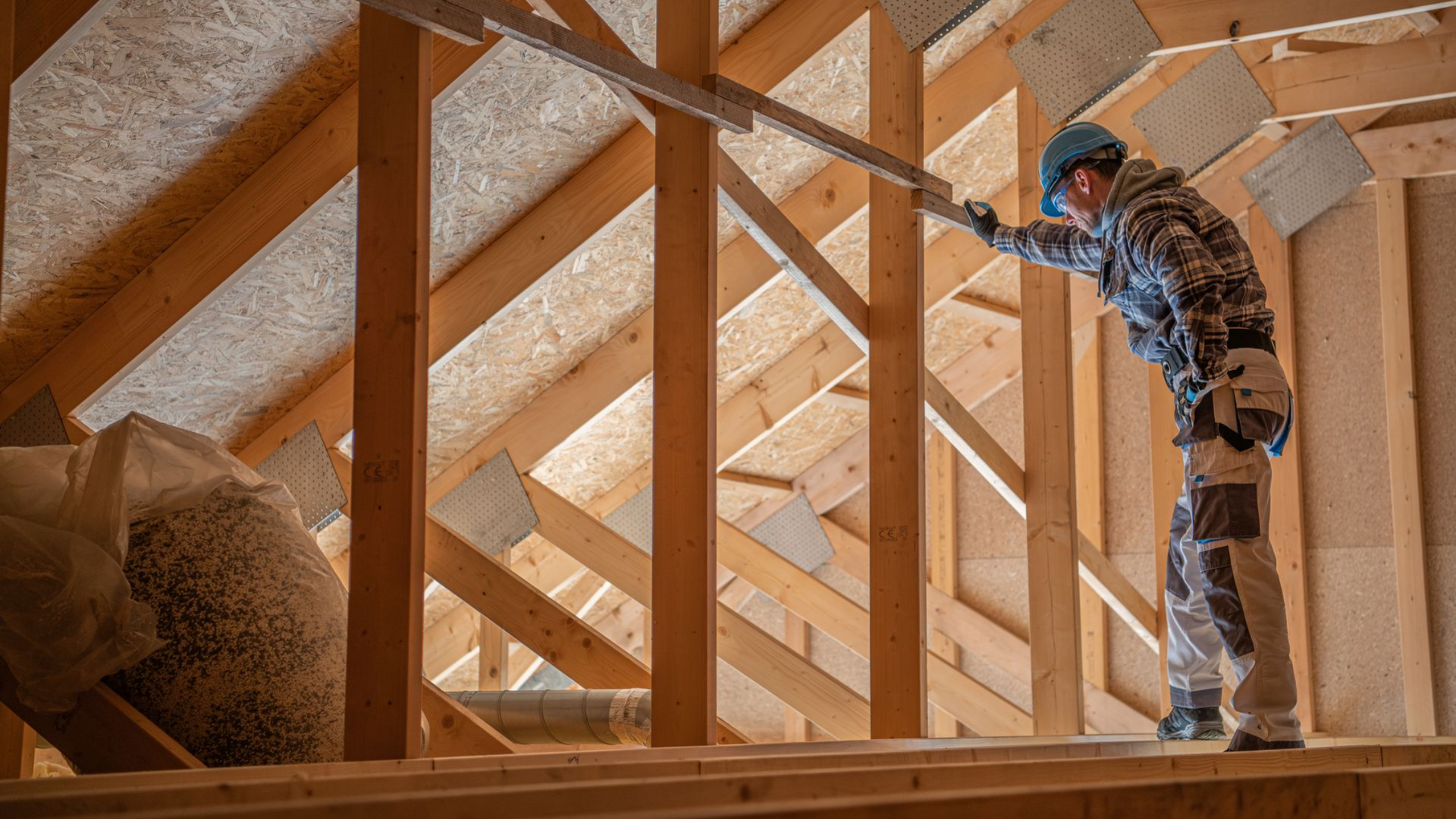 Construction worker in an attic inspecting wooden beams, wearing a hard hat and overalls.