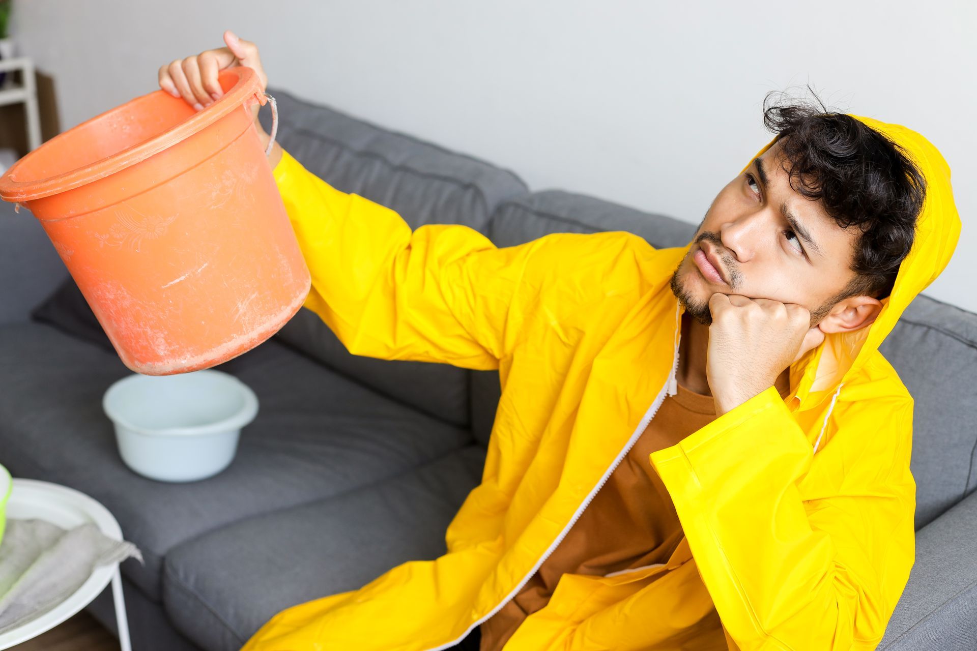 Person in a yellow coat holding an orange bucket over a gray couch while resting their chin on their hand