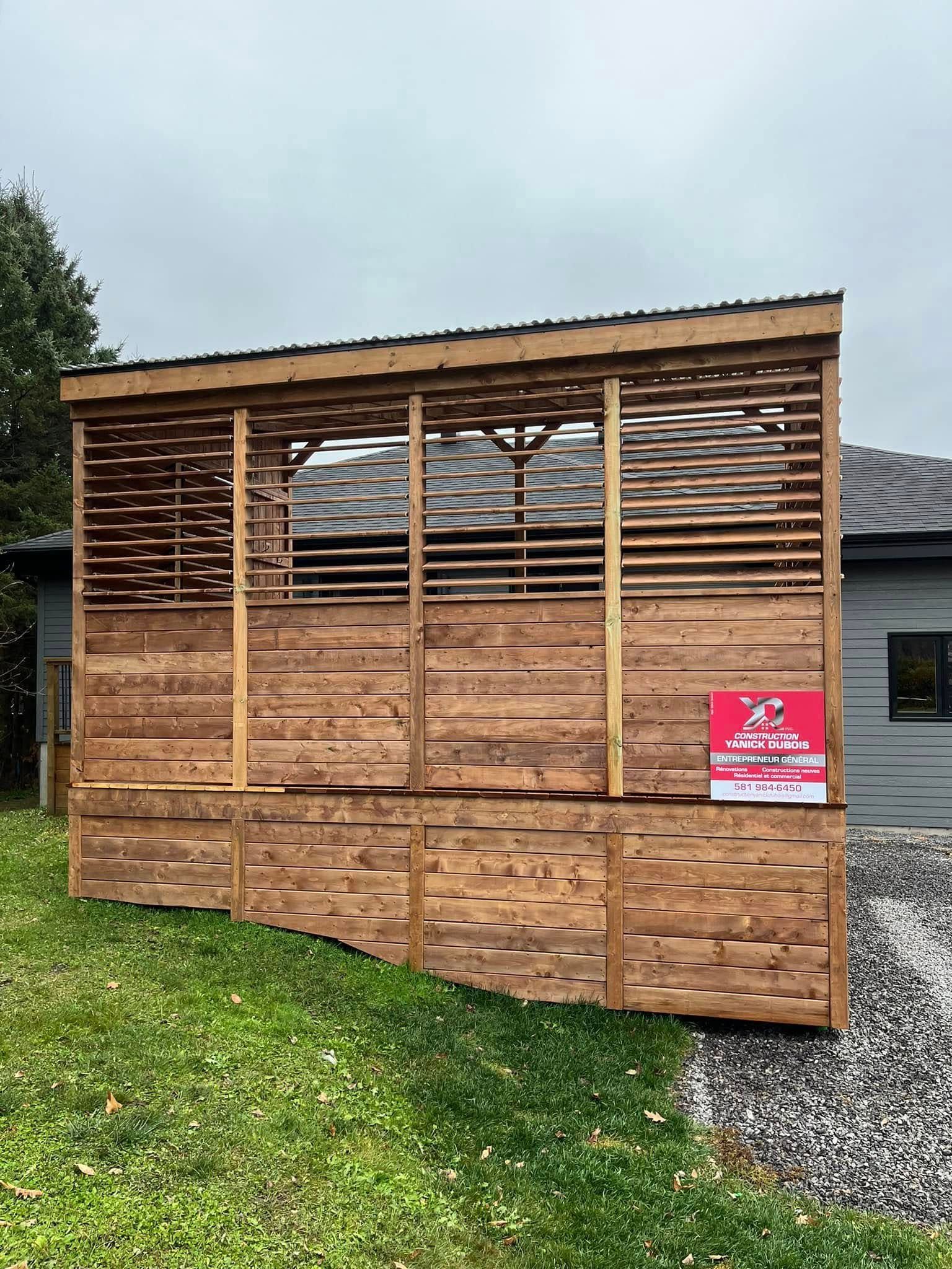 Structure extérieure en bois aux parois à claire-voie, teintée en brun, construite sur une colline herbeuse à côté d'une maison.