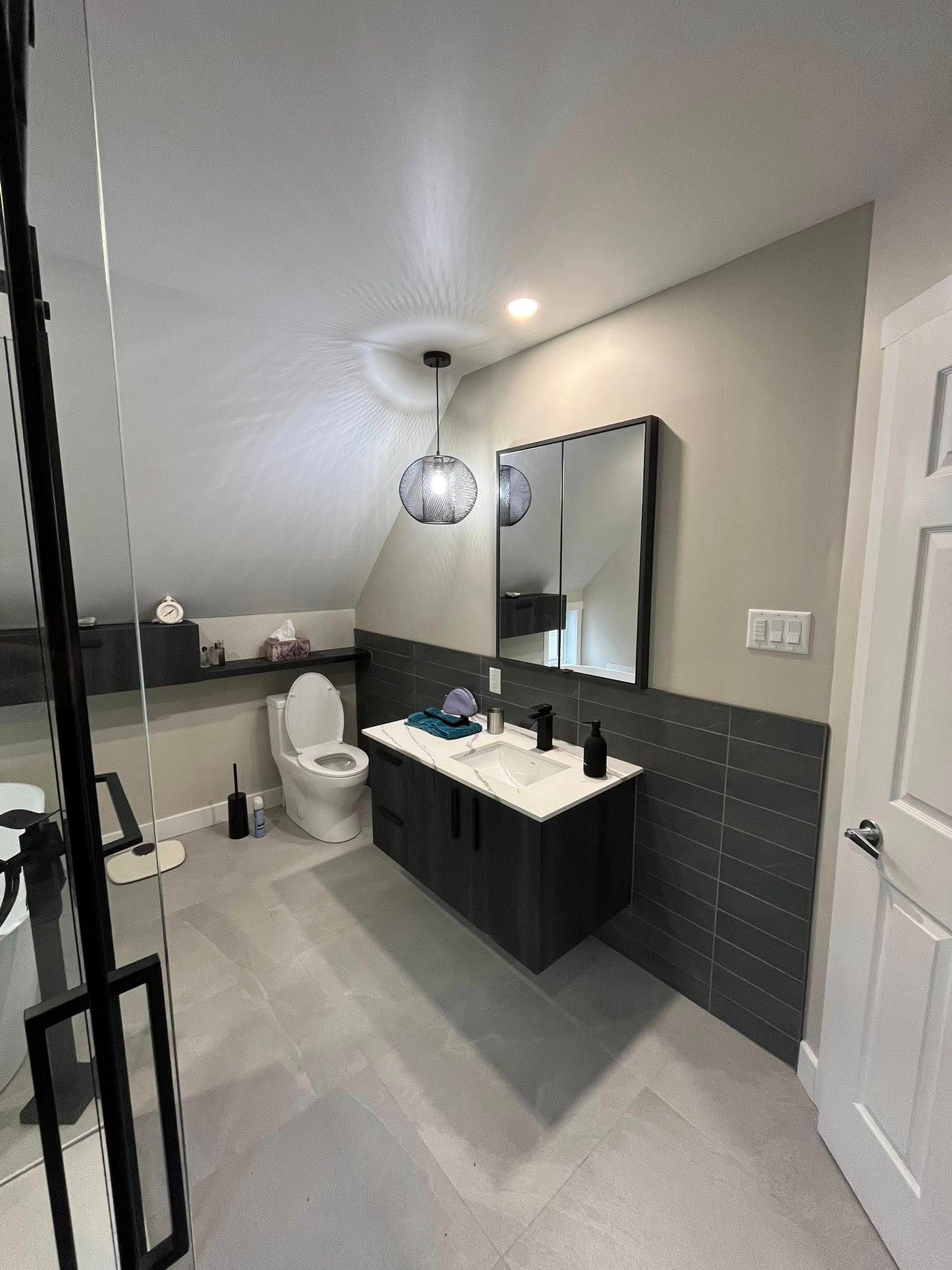 Salle de bains avec meuble-lavabo gris foncé, toilettes et carrelage gris. Murs gris clair, porte blanche et douche en verre.