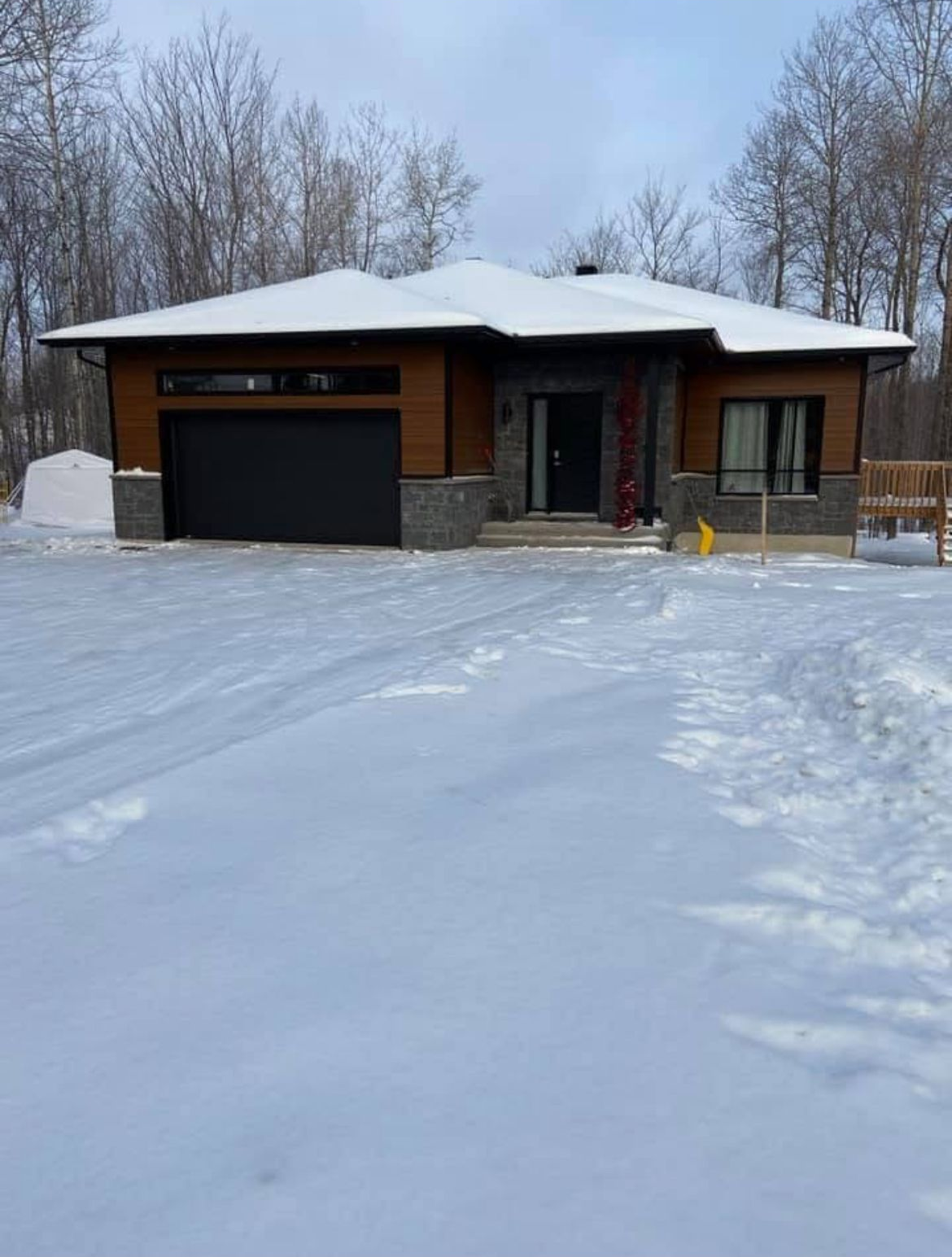 Maison moderne de plain-pied sous la neige ; façade en bois et en pierre ; porte de garage et porte d'entrée noires.