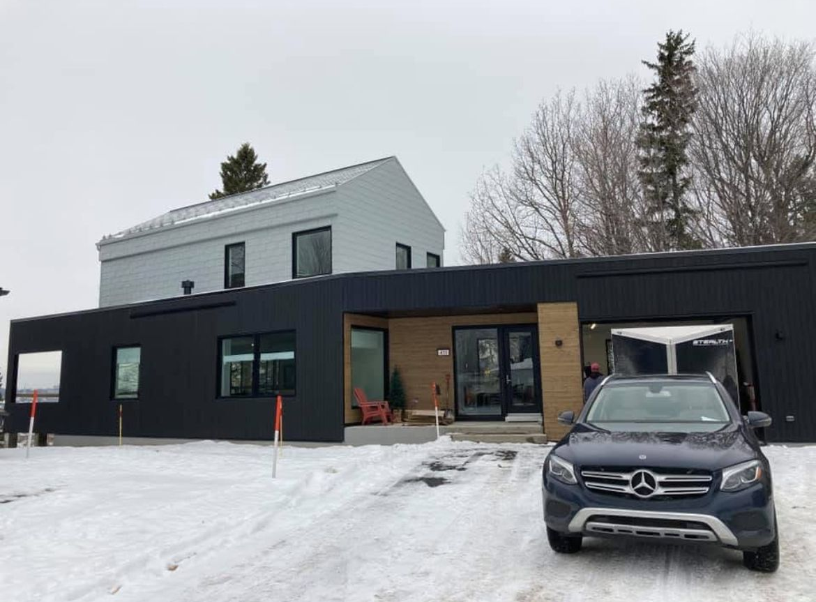 Maison moderne avec un bardage noir et gris, neige au sol et un SUV Mercedes bleu devant.