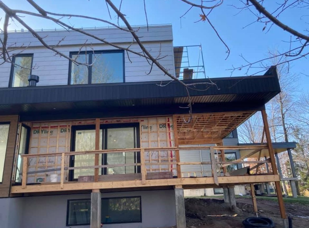Maison en construction ; deux étages avec terrasse inachevée, supports en bois, grandes fenêtres et garnitures de toit noires.