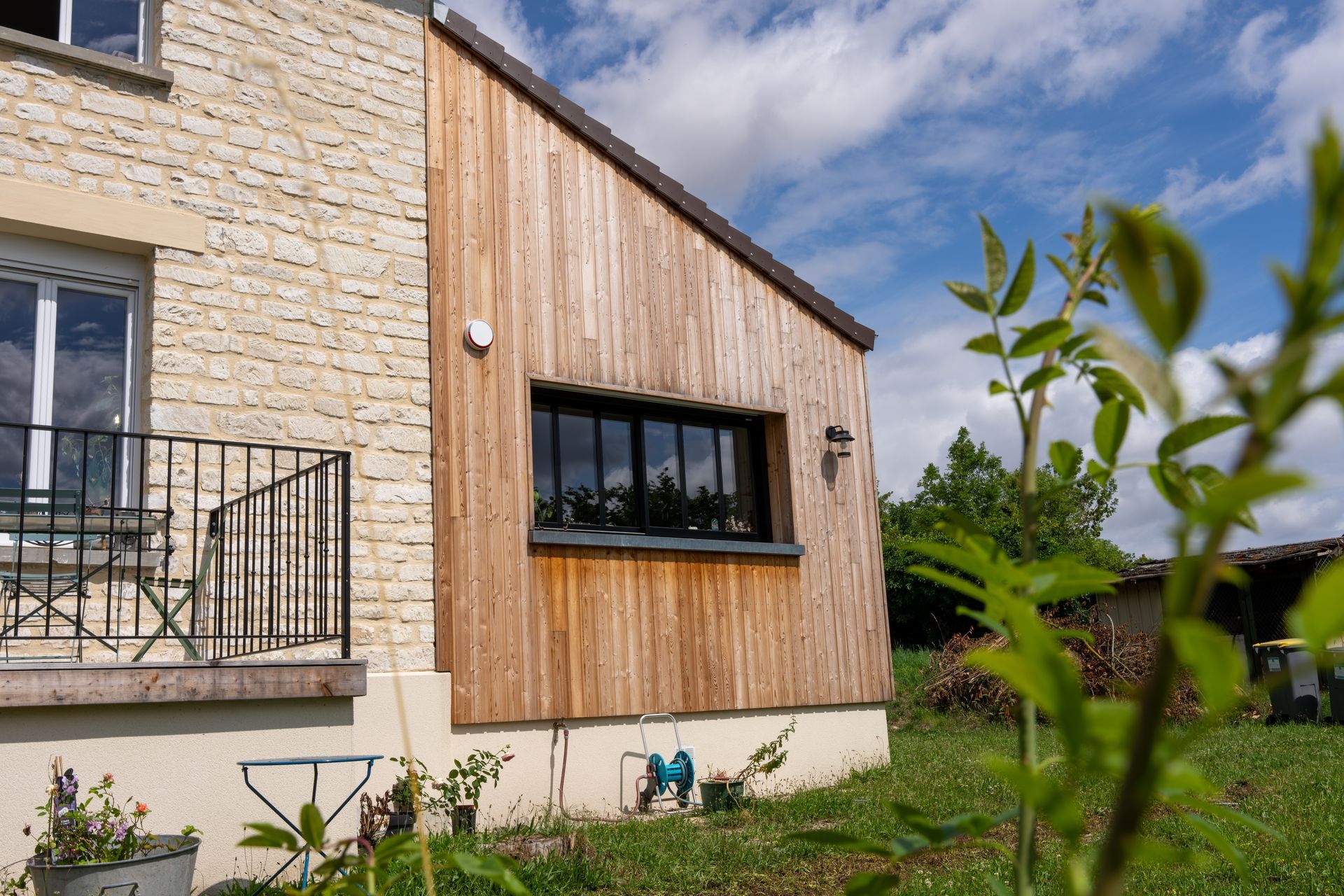 Extérieur de la maison avec revêtement en pierre et en bois, petite fenêtre noire, balcon et jardin.