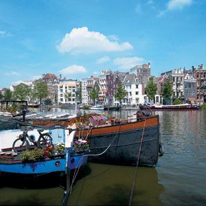 Boats moored in a canal in Amsterdam, Netherlands; buildings in the background under a blue sky.