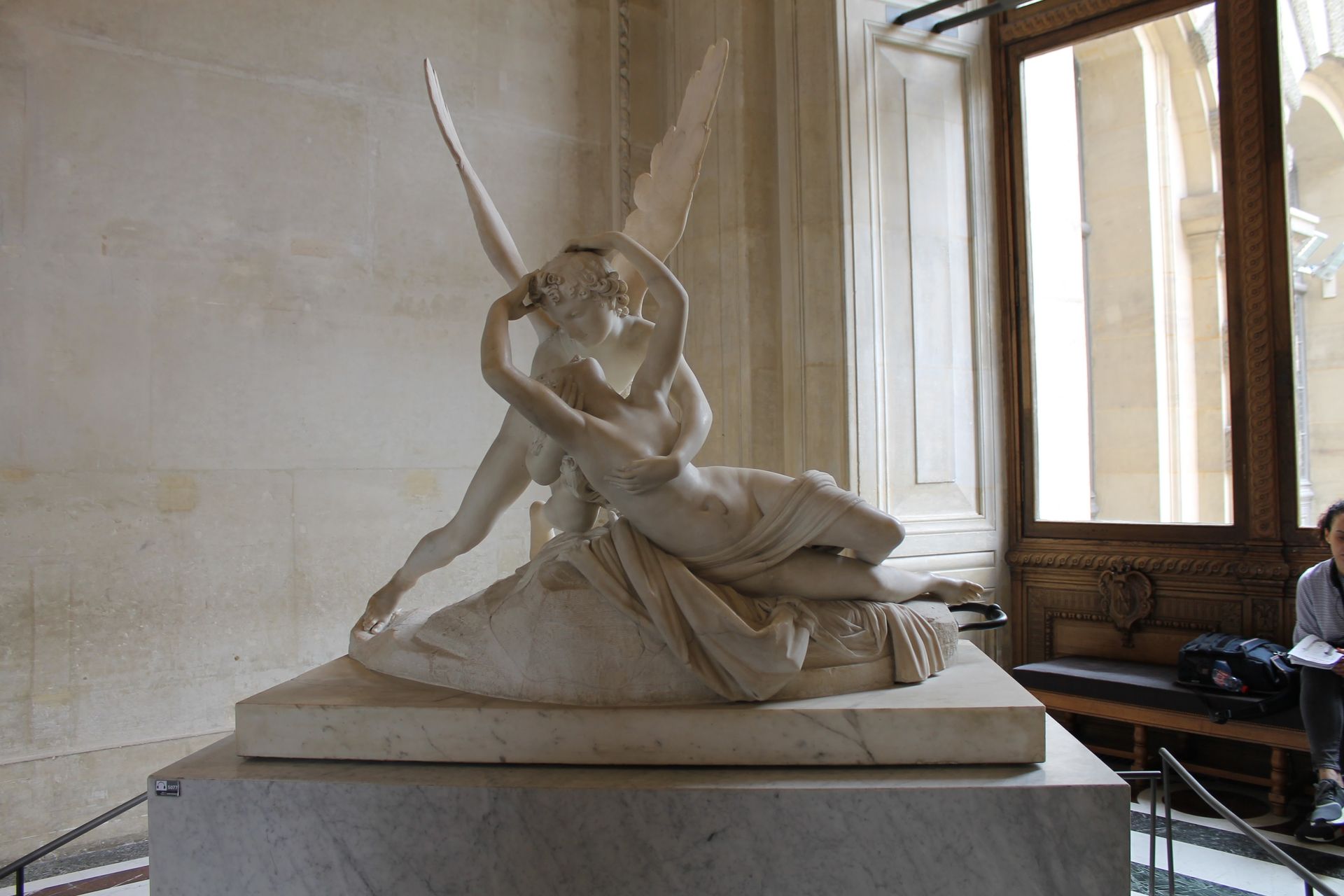 Marble sculpture of Cupid and Psyche embracing. Winged figure leans over reclining woman on a pedestal. Interior with window.