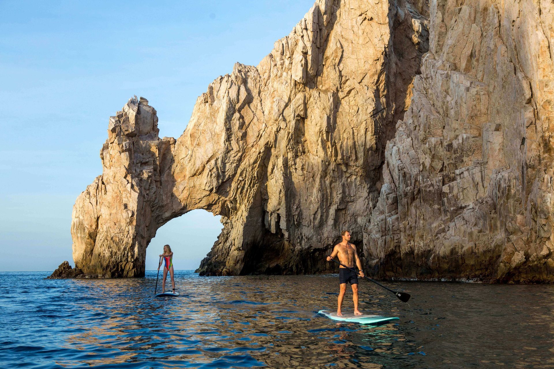 Two people paddleboard near a large rock arch in the ocean.