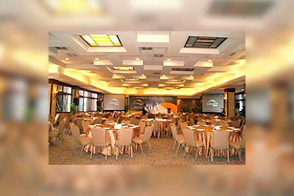 Un amplio salón de banquetes, muy bien iluminado, con mesas redondas cubiertas con manteles beige y sillas, preparado para un evento.