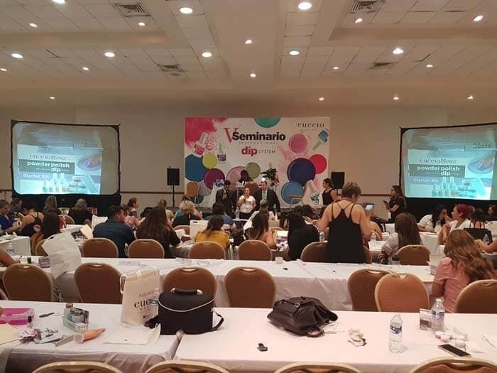 El público se sienta en mesas en una gran sala de conferencias frente a un escenario con un colorido telón de fondo para seminarios y dos pantallas.