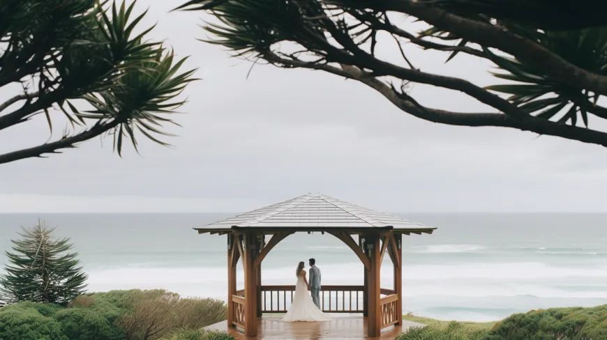 A bride and groom are standing under a gazebo overlooking the ocean.