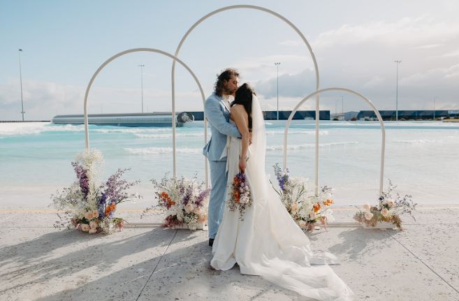 A bride and groom are kissing under a white arch on the beach.