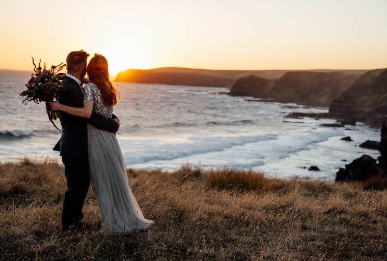 A bride and groom are kissing in front of the ocean at sunset.