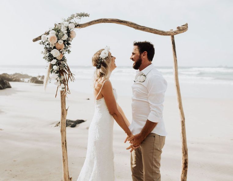 A bride and groom are holding hands on the beach under a wooden arch.