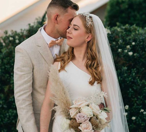 A bride and groom are posing for a picture and the groom is kissing the bride on the cheek.
