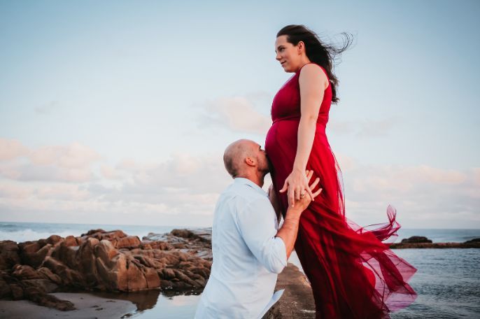 A man is kissing a pregnant woman 's belly on the beach.