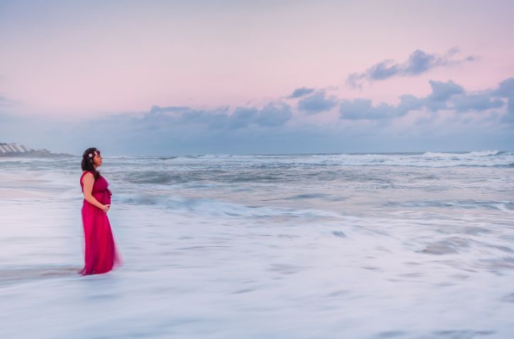 A pregnant woman in a red dress is standing on the beach looking at the ocean.