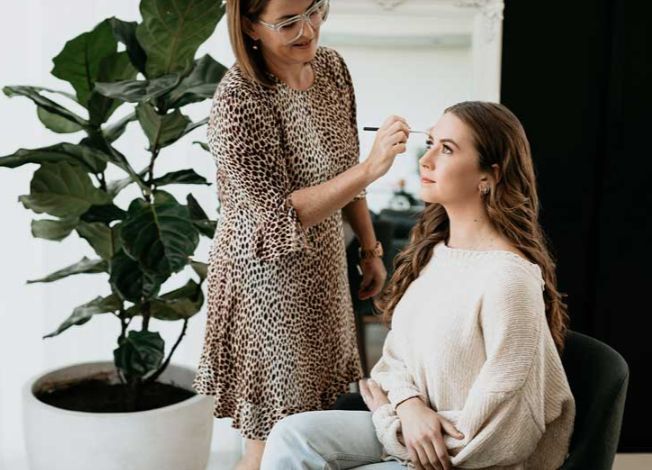 A woman is applying makeup to a woman sitting in a chair.