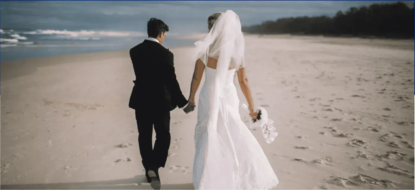 A bride and groom are walking on the beach holding hands.
