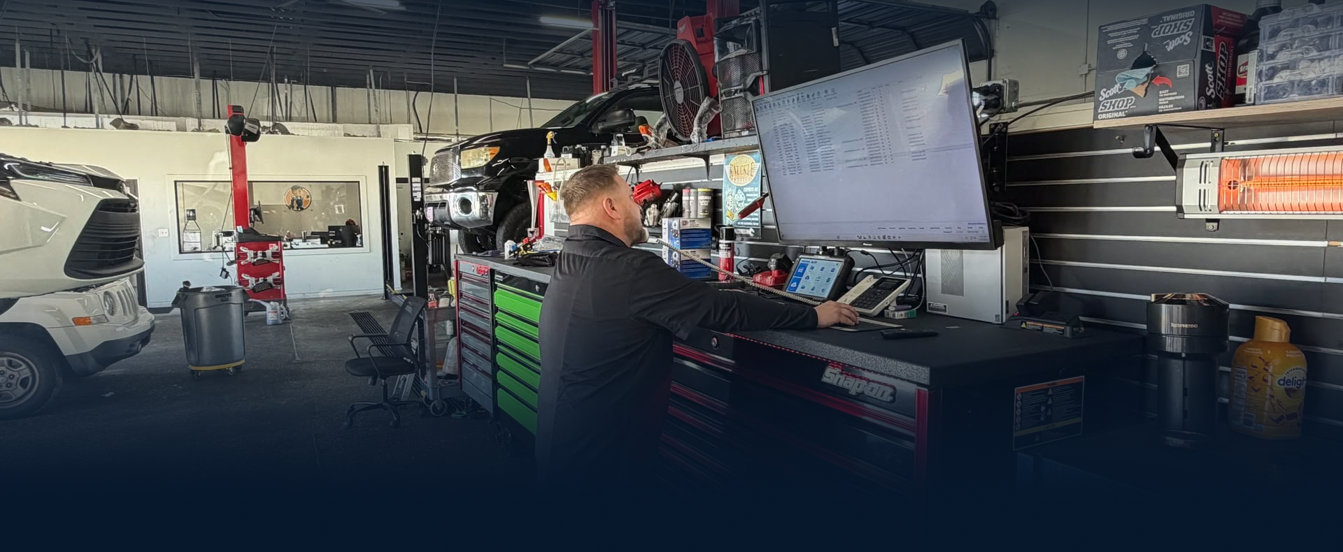 Mechanic working at a garage workstation with computers, tools, and a car in the background | Dunedin Total Car Care