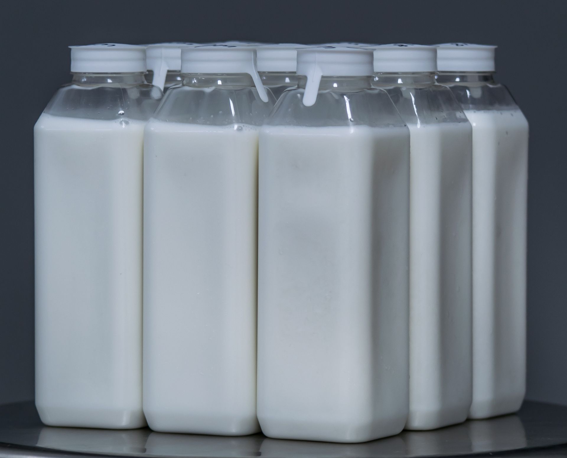 Five clear plastic milk bottles filled with white liquid, arranged side by side on a dark surface.