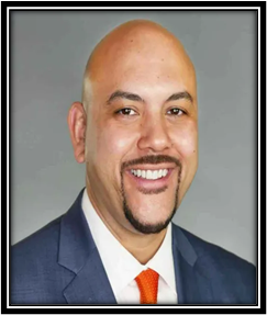 Man in blue suit, orange tie, bald head, goatee, smiling.