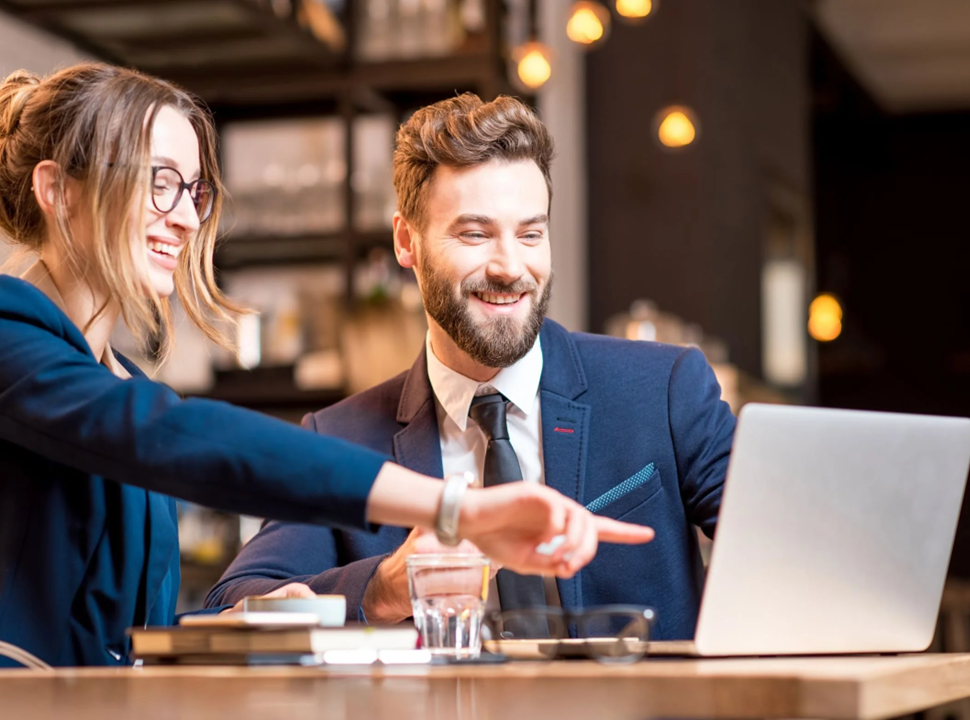 Two people in business attire look at a laptop, smiling, in a cafe. The person on the right points.
