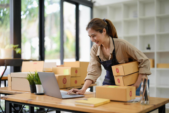 Woman in apron smiles while using laptop, holding packages, preparing online orders.