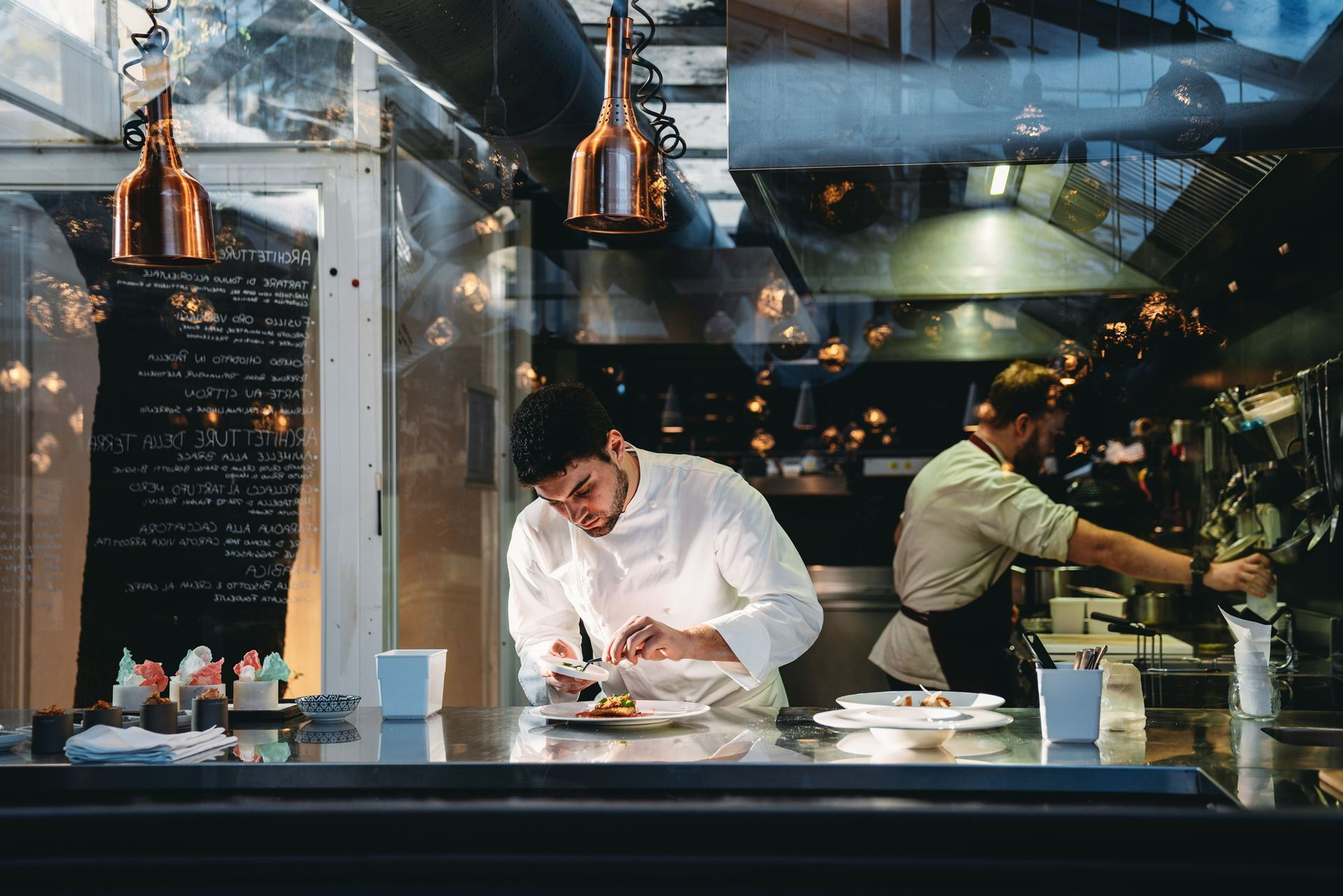 Chefs in a restaurant kitchen, one preparing a dish at a stainless steel counter.