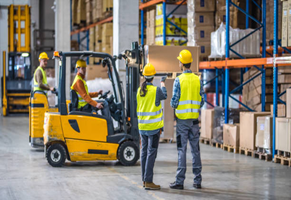 Workers in a warehouse with forklifts and shelves of boxes. They are wearing safety vests and hard hats.