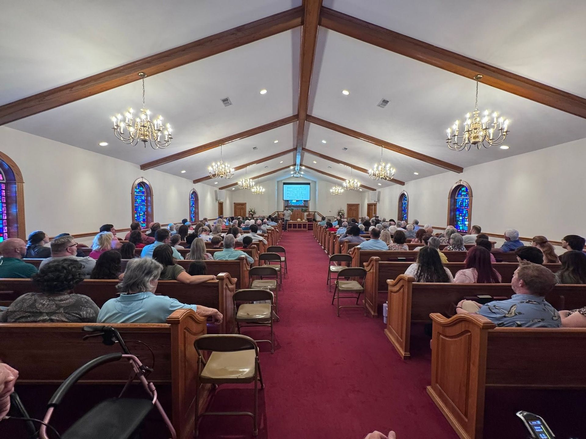 Church sanctuary with congregants seated in pews, red carpet aisle, chandeliers, and a pulpit at the front