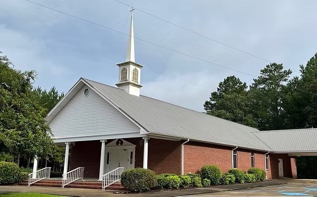 Brick church with white steeple, red doors, and gray roof surrounded by trees