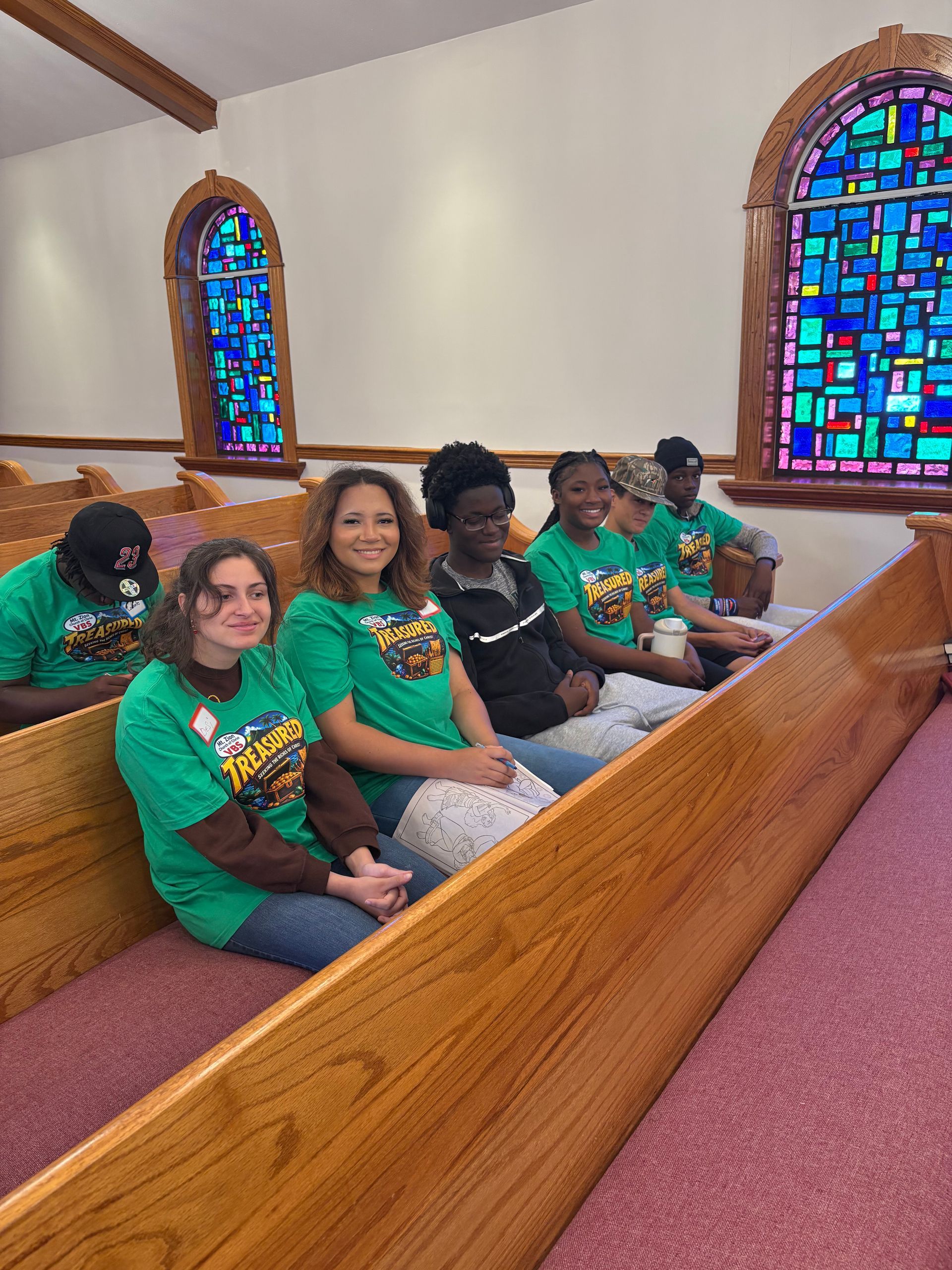 Six people in green shirts seated in a church pew beside colorful stained-glass windows.