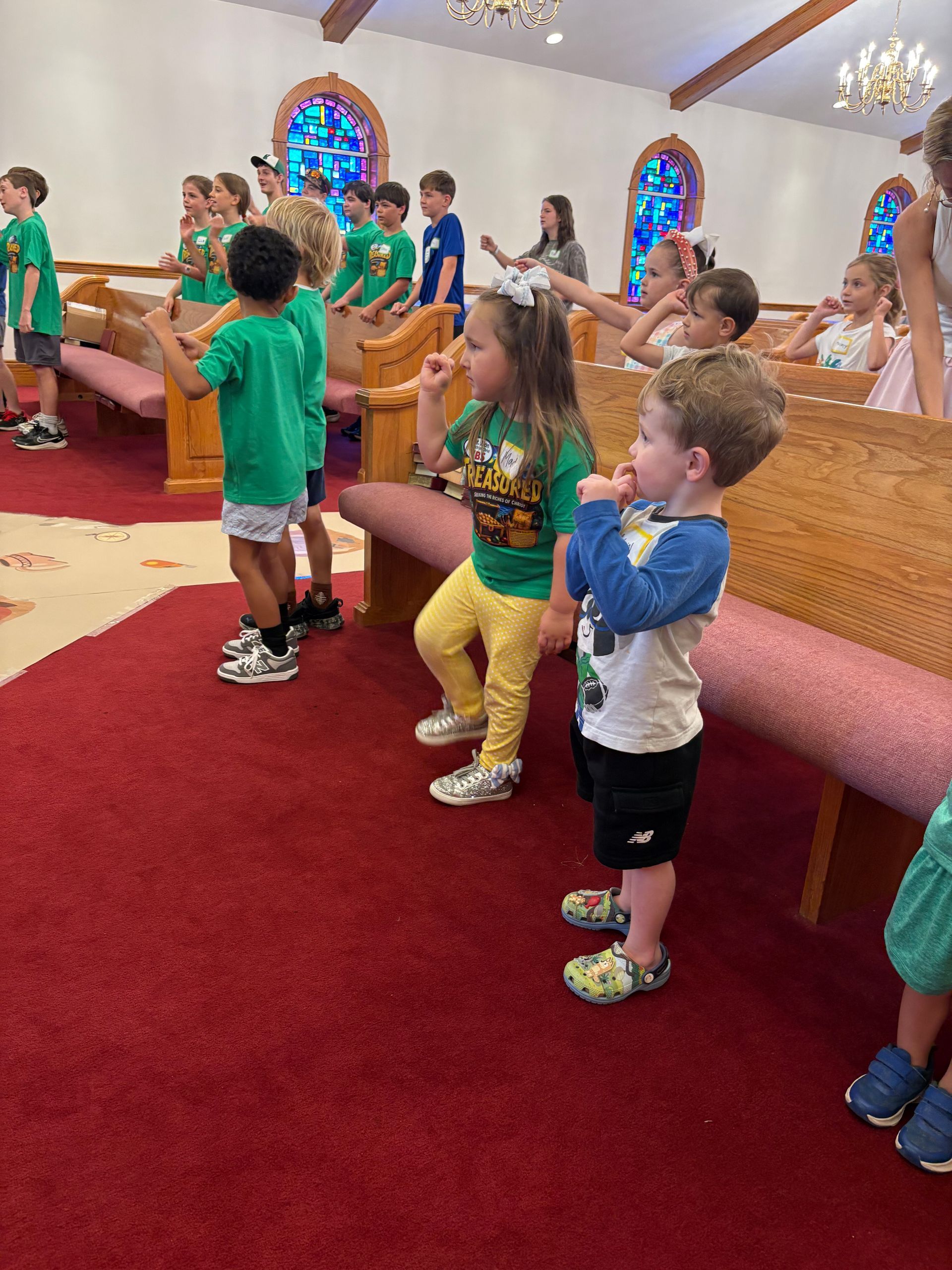 Children in a church sanctuary standing and singing on red carpeted steps with stained-glass windows behind them
