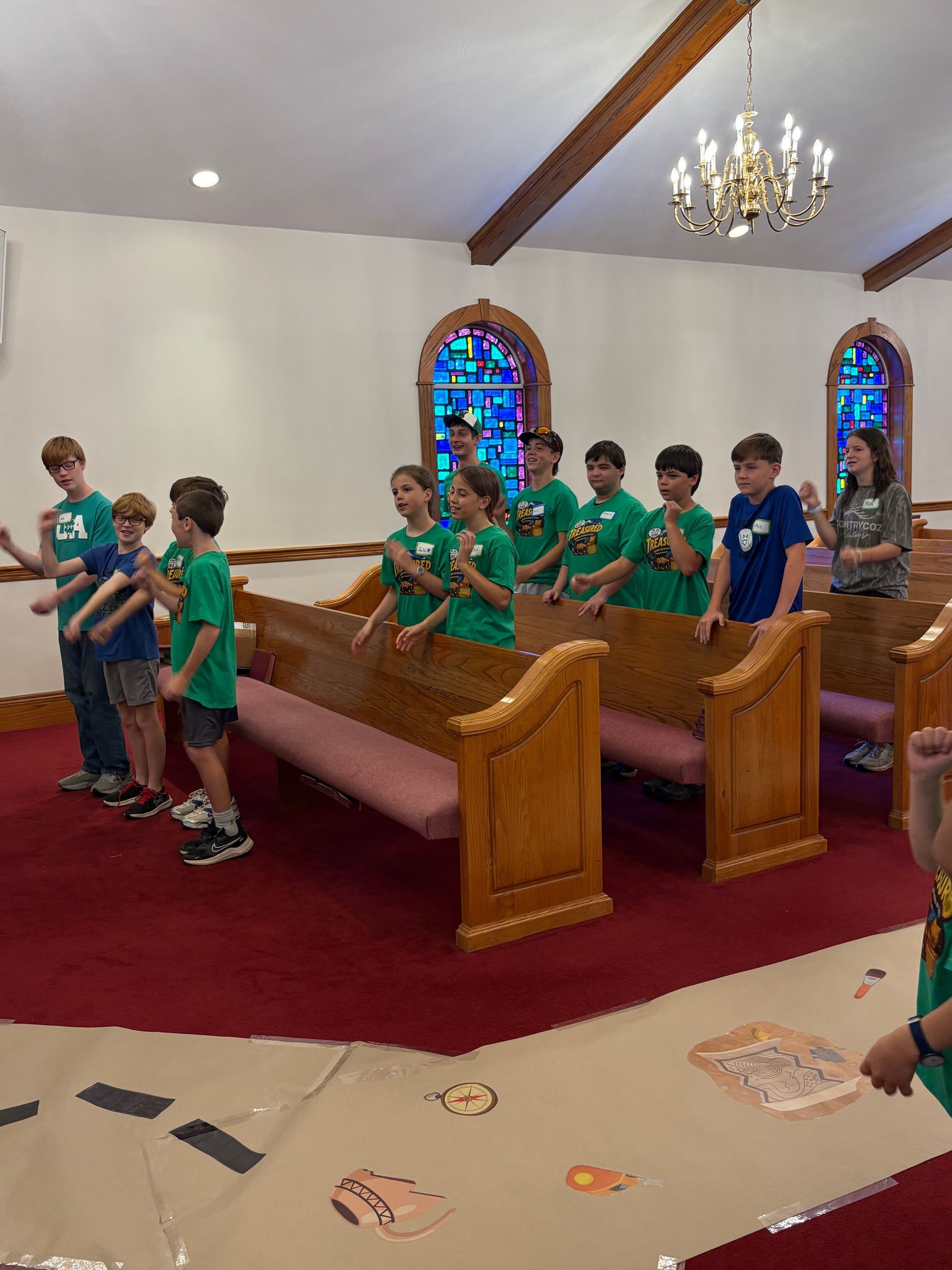 Children in green shirts stand in wooden church pews during a service, with stained glass and a chandelier.