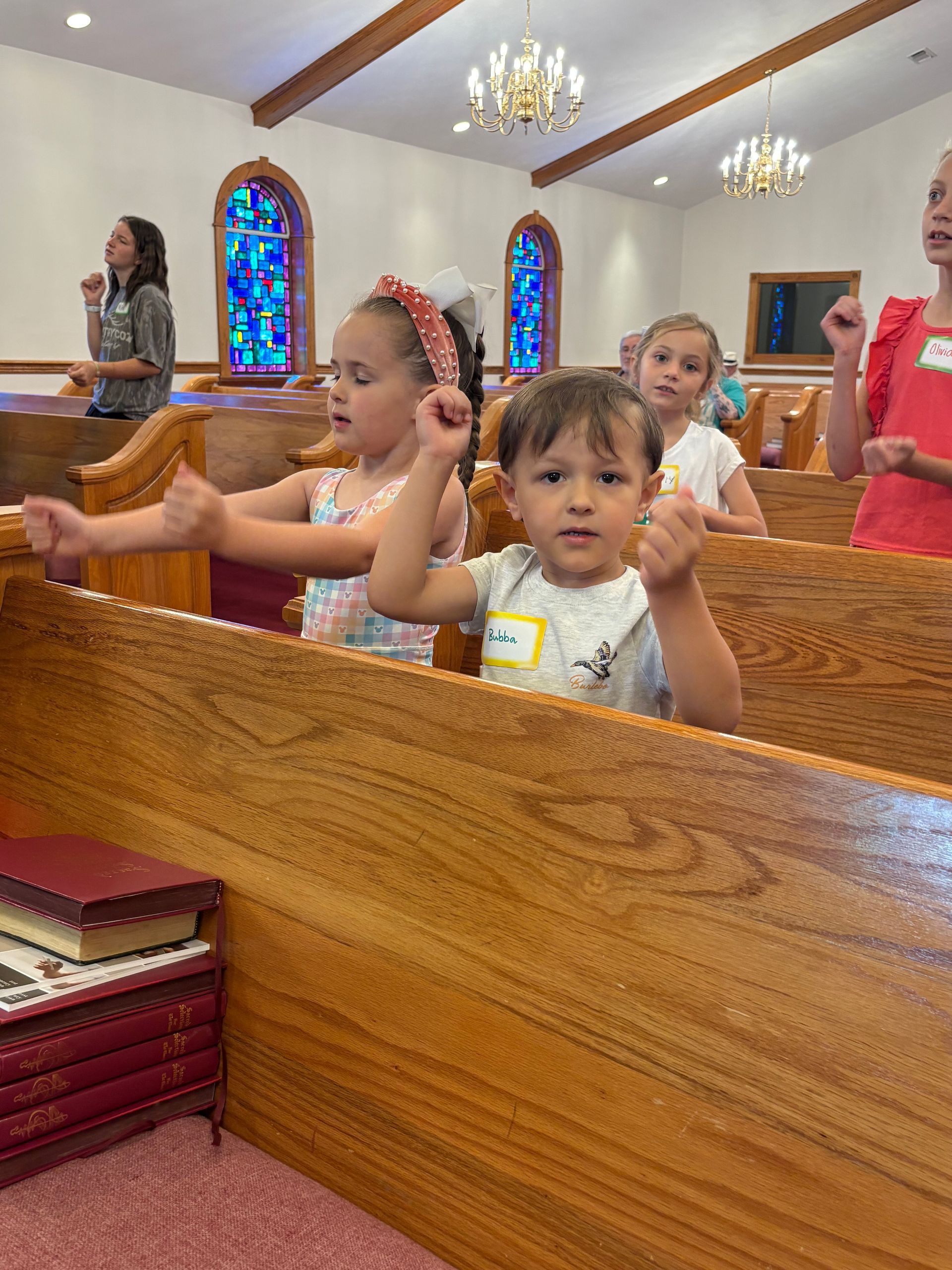 Children sitting in church pews during a service, with stained-glass windows in the background.