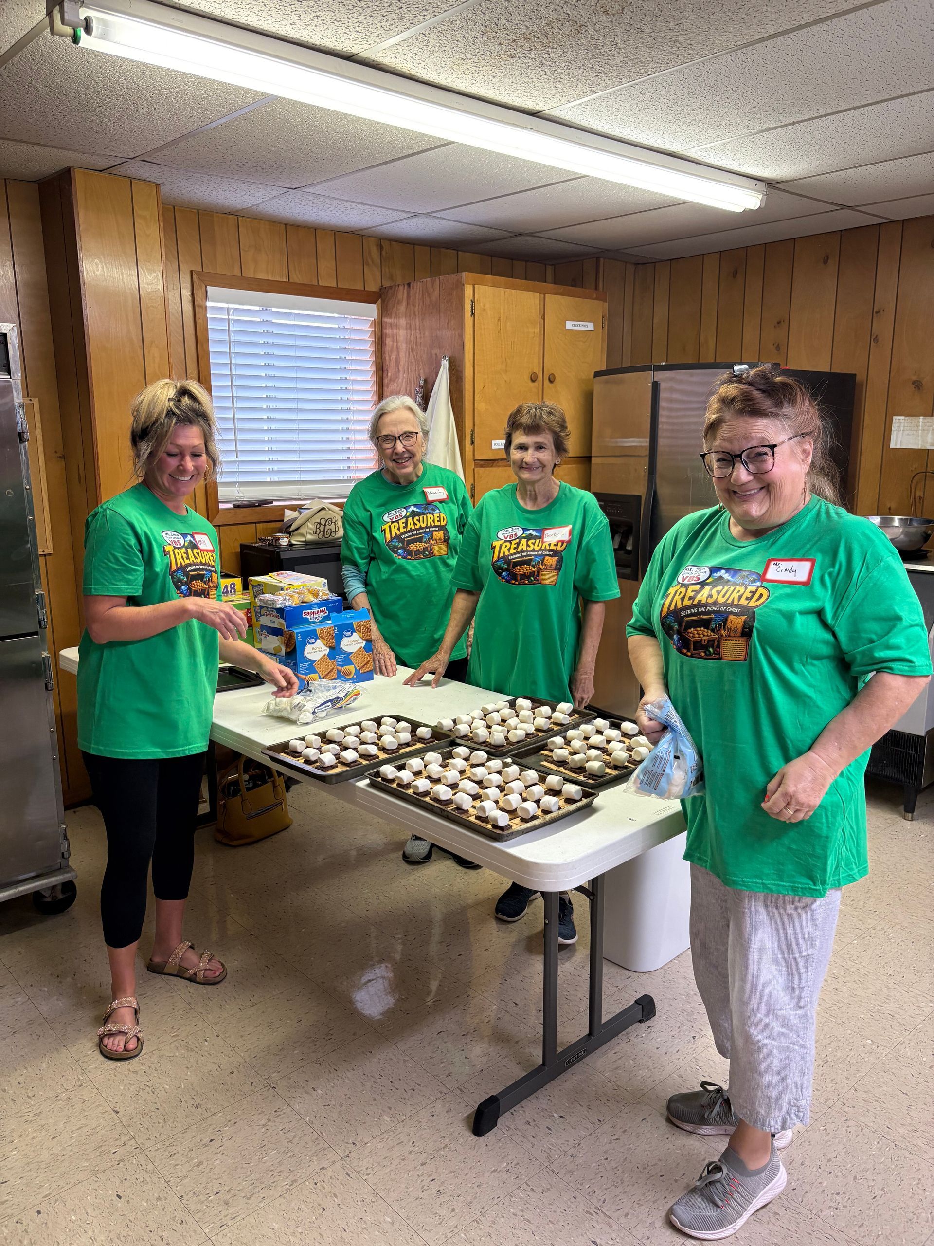 Four people in green shirts stand around a table of baked goods in a kitchen.
