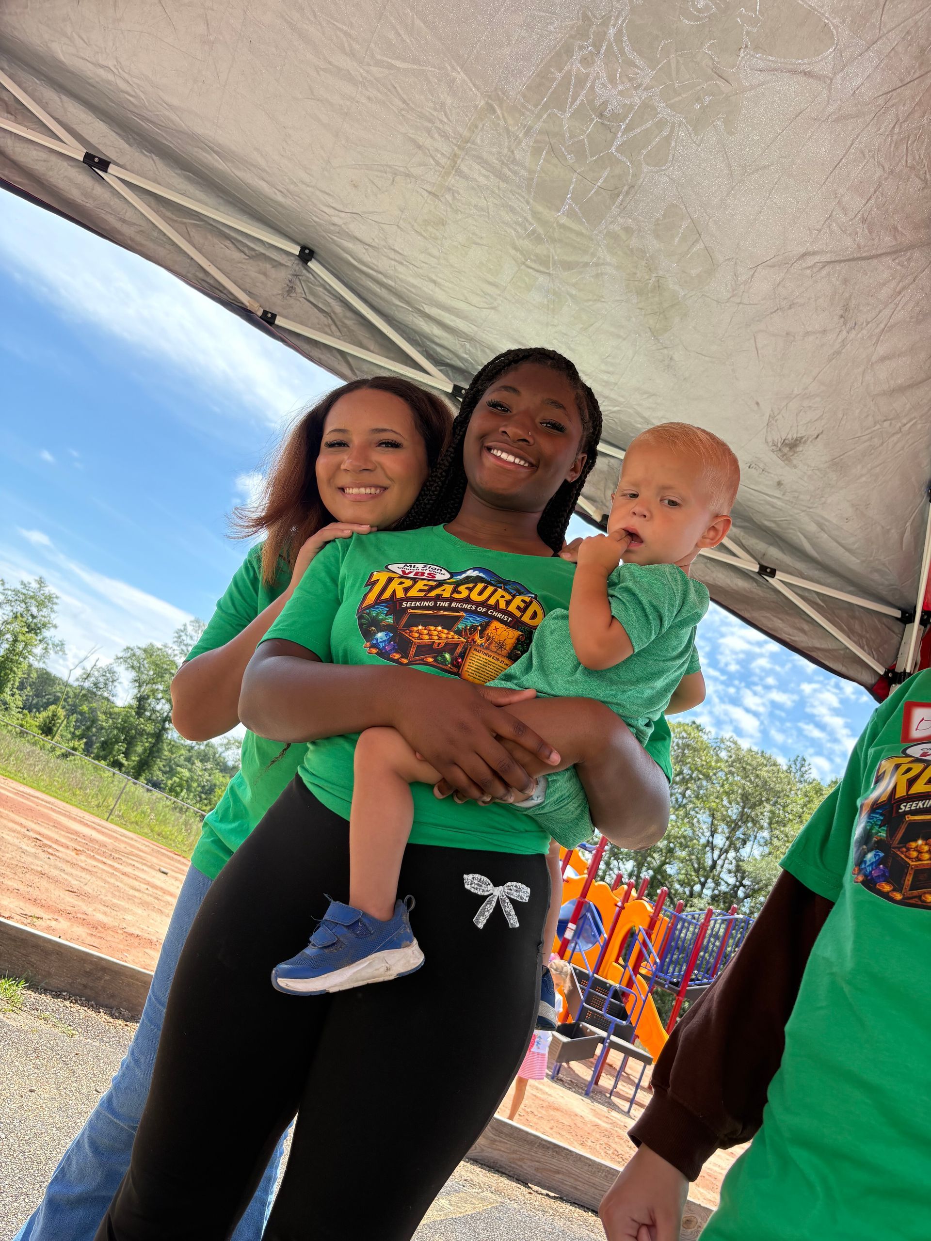 Three people in green shirts holding a baby under a tent at an outdoor event.