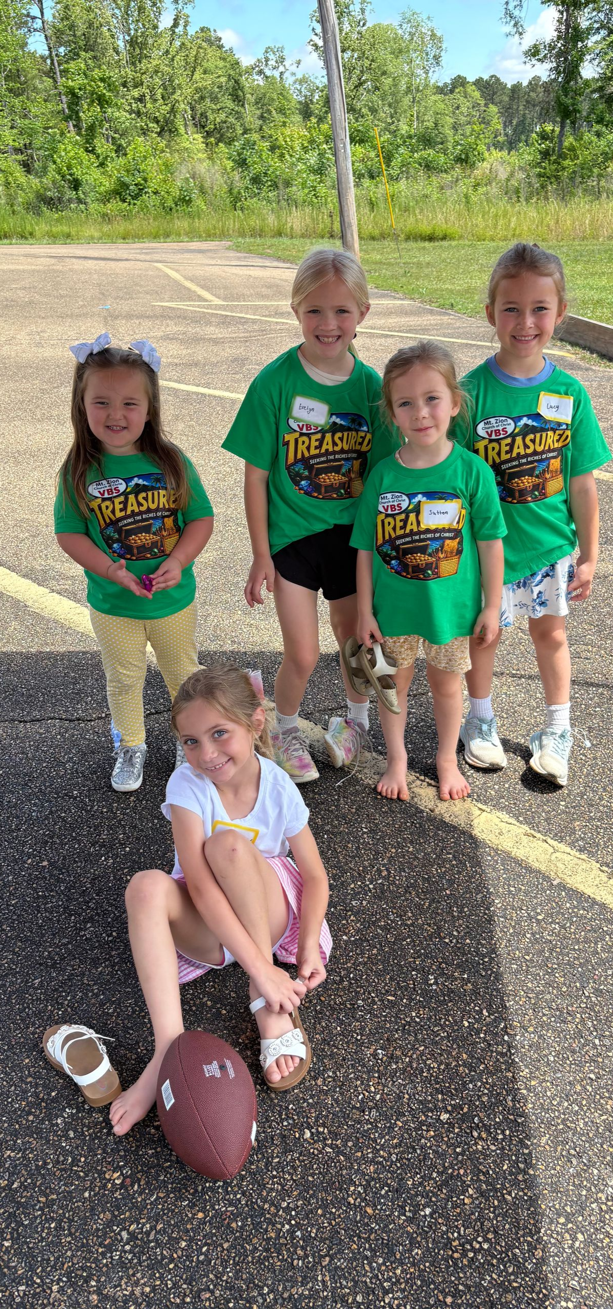 Five children in matching green shirts stand outdoors by a football; one child sits on the ground.