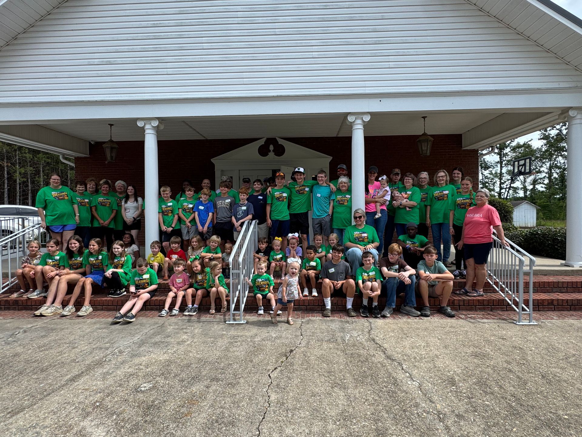 Large group posing on porch steps under a pavilion, many in green shirts with a few adults in red.