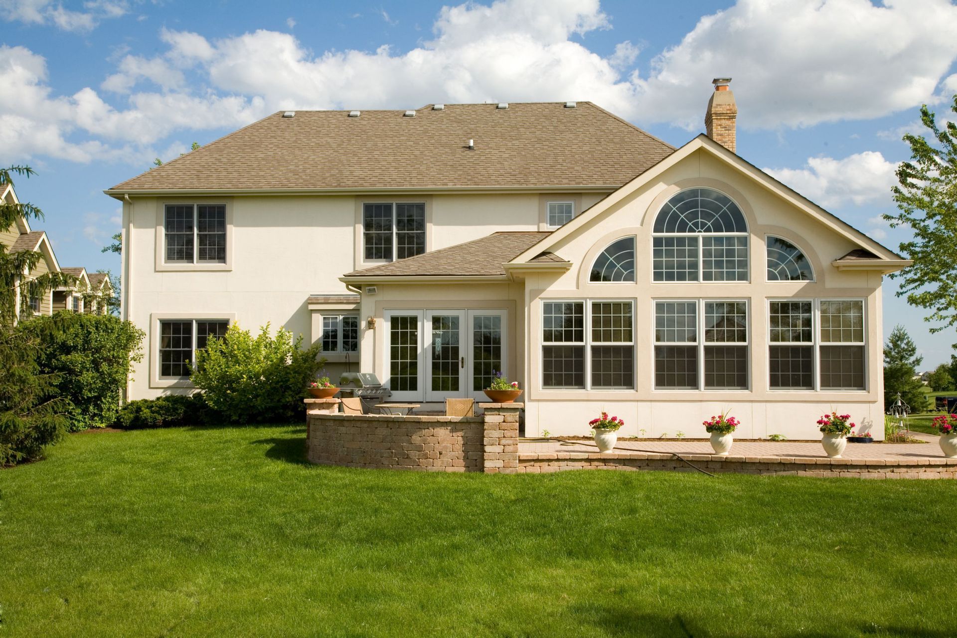 Two-story beige house with a sunroom addition and stone patio, surrounded by a lush green lawn under a blue sky.