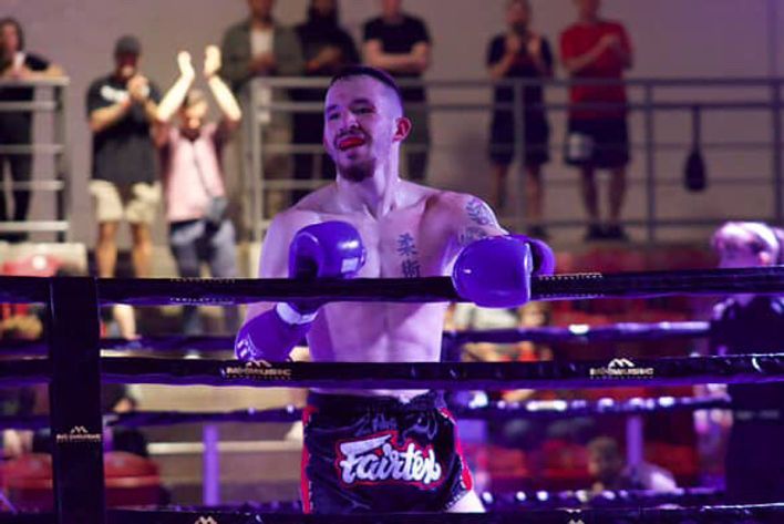 Boxer in purple gloves and black trunks stands in the ring under bright lights.