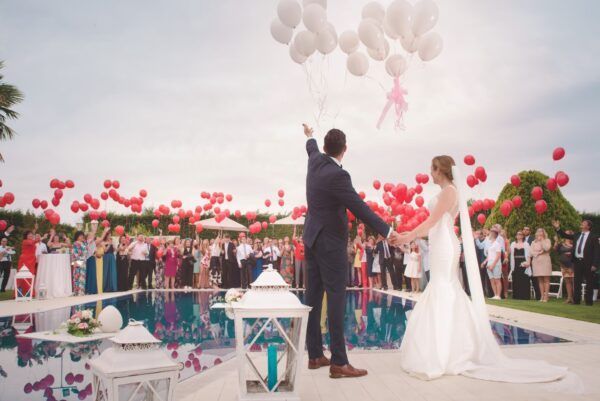 Bride and groom releasing red and white balloons at outdoor wedding near pool. Guests watch.
