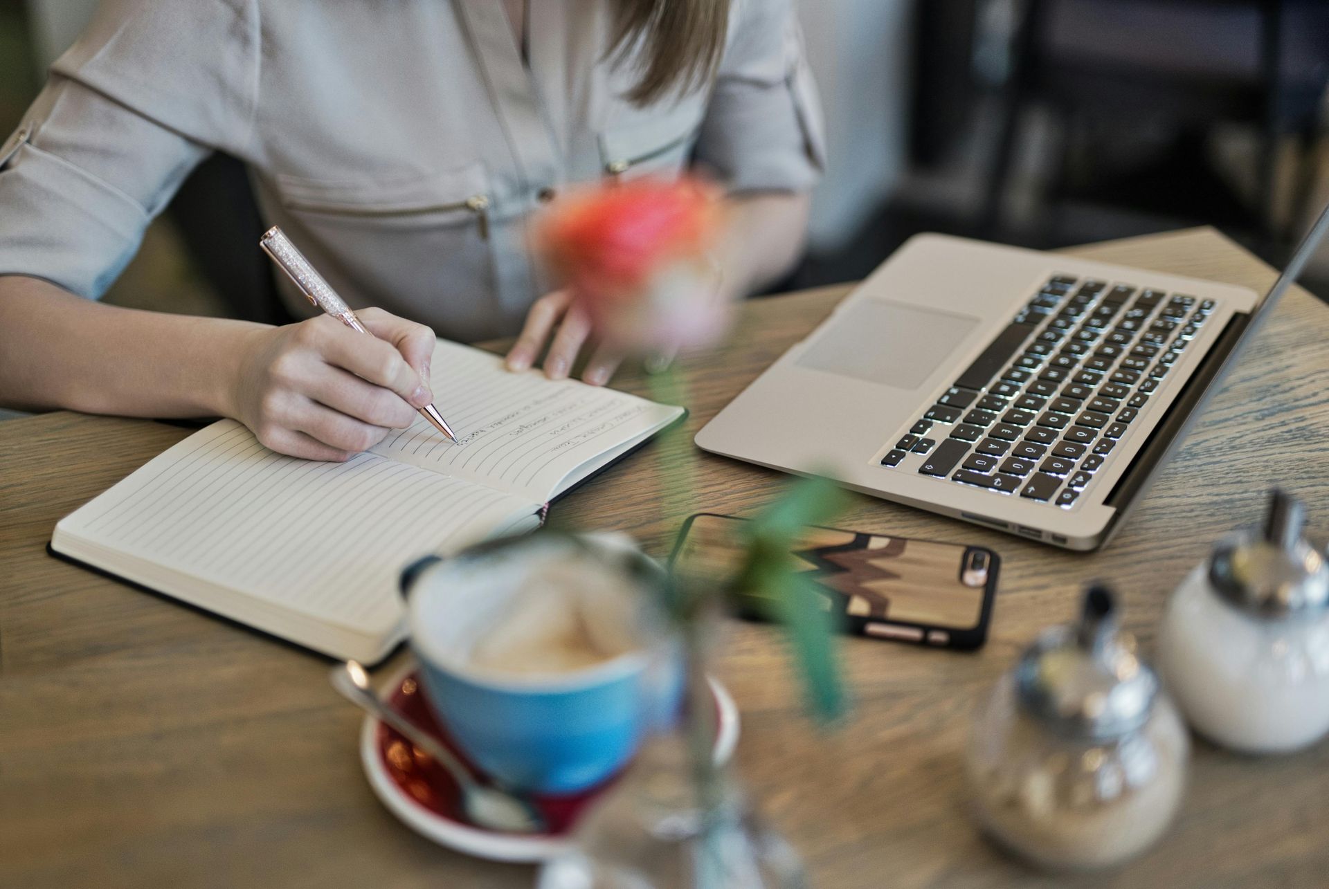 Woman writing in notebook at cafe table with laptop, coffee, and flowers.