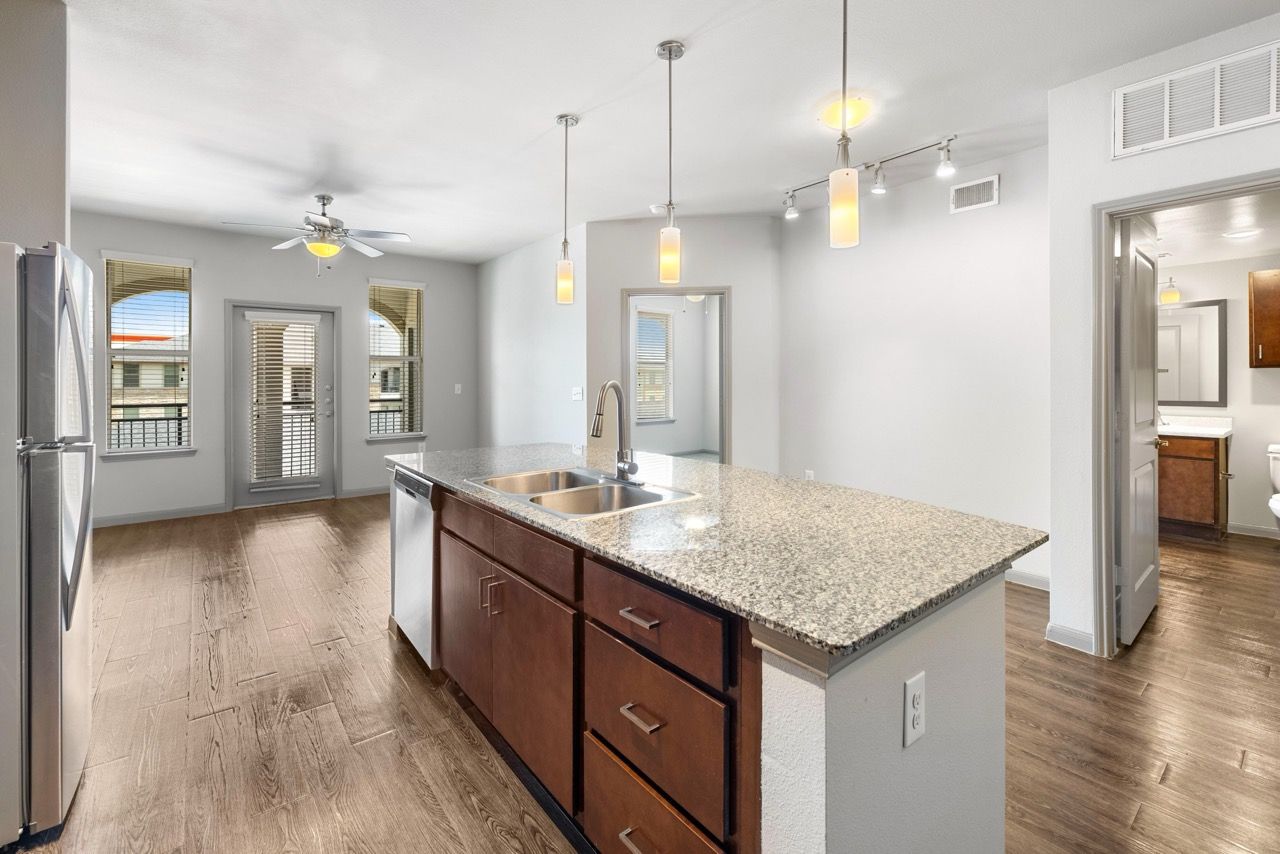 Open-concept kitchen with granite island, stainless steel appliances, pendant lighting, and hardwood floors in an apartment.