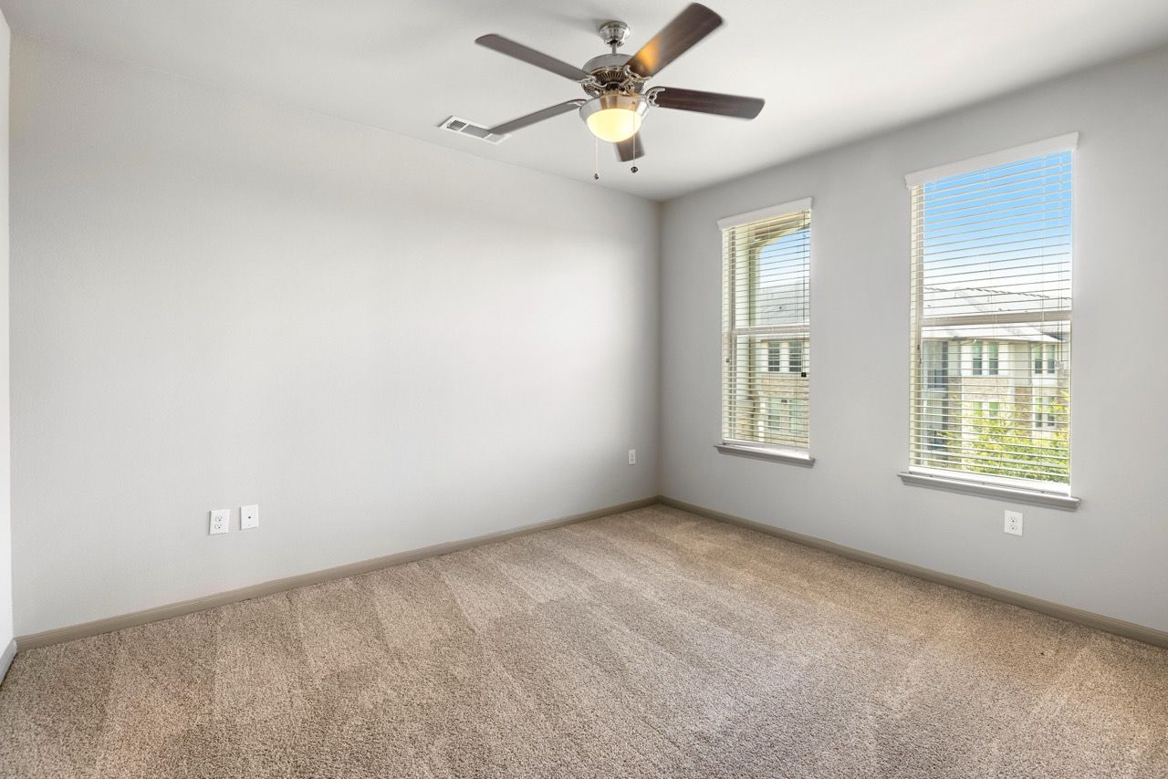 Empty bedroom with beige carpeting, light gray walls, two windows with blinds, and a ceiling fan.